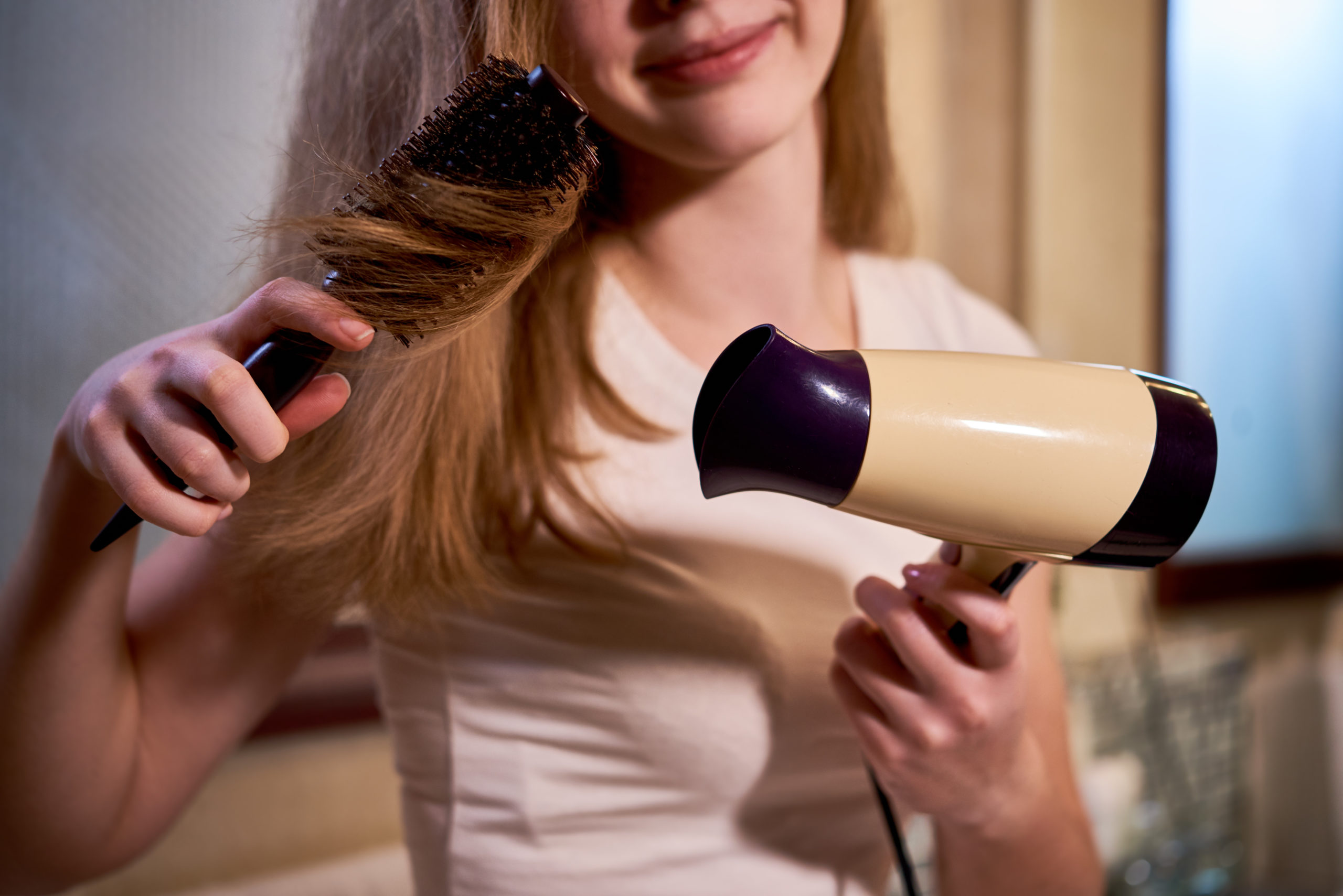 Young girl standing and making a hairstyle with brush and hairdryer in the bathroom at home